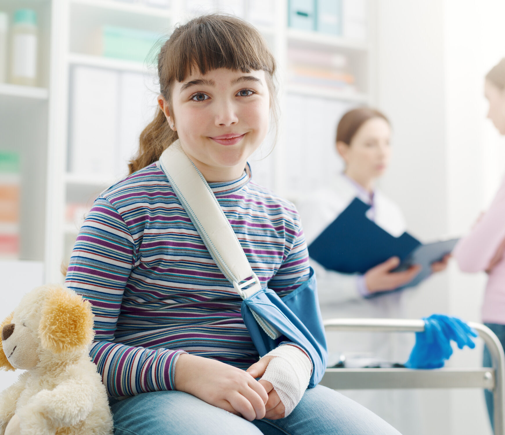 Lächelndes Mädchen mit Verband und Armspange mit Teddybär daneben sitzend